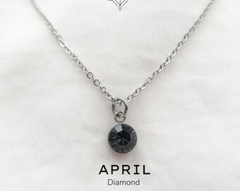 Puede incluir: Un collar de cadena de plata con un pequeño colgante de cristal negro. El colgante es redondo y tiene una superficie facetada. El texto "APRIL Diamond" está escrito debajo del colgante.