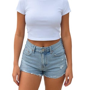 May include: A white, short-sleeved crop top paired with light blue denim shorts. The shorts have a distressed look with frayed edges. The outfit is simple and casual, suitable for warm weather.