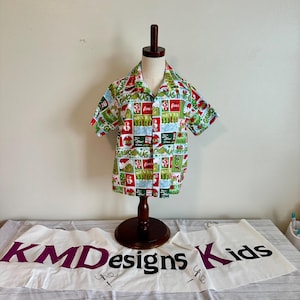 May include: A short-sleeved button-down shirt with a festive Grinch-themed pattern. The shirt features a white background with red, green, and blue squares, each containing Grinch-related imagery and text. The shirt is displayed on a wooden mannequin.