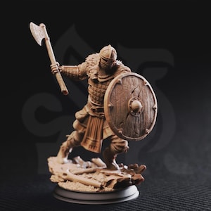 Puede incluir: Figura marrón detallada de un guerrero vikingo en posición de batalla. El guerrero lleva un casco, armadura y una capa con ribete de piel, empuñando un hacha y un escudo redondo. La figura se alza sobre una base texturizada, sugiriendo un campo de batalla.