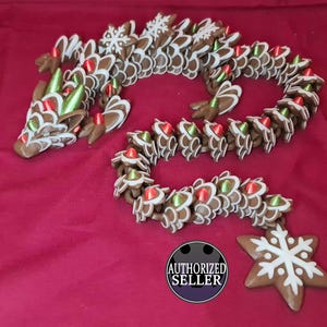 Puede incluir: Un dragón de jengibre marrón y blanco con detalles rojos y verdes. El dragón está hecho de múltiples piezas de jengibre y tiene un dije en forma de copo de nieve. Sobre una superficie roja.