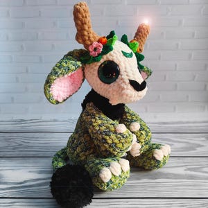 Könnte beinhalten: Ein handgefertigtes Häkeltier mit großen, glitzernden Augen, braunen Geweihen und einem Blumenkranz. Das Plüschtier hat einen grün-weiß gemusterten Körper, rosa Innenohren und einen schwarzen Kragen. Die Kreatur sitzt auf einer Holzoberfläche.