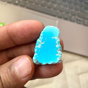 May include: A polished turquoise gemstone, displaying a vivid blue colour and a textured, uneven edge. The stone is held in a hand, highlighting its natural form. The gemstone is a striking example of natural turquoise.