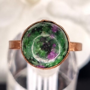 May include: A copper ring featuring a green and pink gemstone. The gemstone is set in a bezel setting and has a smooth, polished finish.