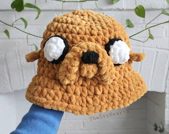 May include: A crocheted brown bucket hat with a cartoon dog face. The hat has white eyes, a black nose, and black eyebrows.