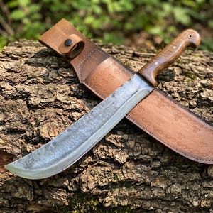 May include: A large, curved blade knife with a wooden handle and a leather sheath. The blade has a textured finish. The sheath is brown leather with decorative embossed patterns. The knife is resting on a textured wooden surface.