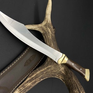 May include: A fixed-blade knife with a curved silver steel blade, a dark brown wooden handle, and polished brass bolster and pommel. The knife is accompanied by a brown leather sheath featuring decorative stitching and an embossed pattern.