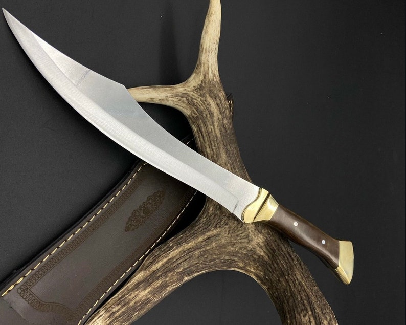 May include: A fixed-blade knife with a curved silver steel blade, a dark brown wooden handle, and polished brass bolster and pommel. The knife is accompanied by a brown leather sheath featuring decorative stitching and an embossed pattern.