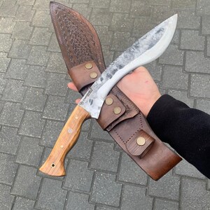 May include: A large, curved knife with a wooden handle and a textured, silver blade. The knife is in a brown leather sheath with decorative stitching and brass-colored rivets. The knife is designed for outdoor use.