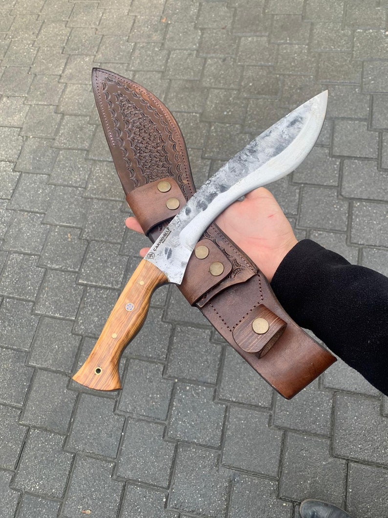 May include: A large, curved knife with a wooden handle and a textured, silver blade. The knife is in a brown leather sheath with decorative stitching and brass-colored rivets. The knife is designed for outdoor use.