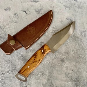 K&ouml;nnte beinhalten: Eine braune Lederscheide mit einem dekorativen Design und ein Messer mit Holzgriff und silberner Klinge.