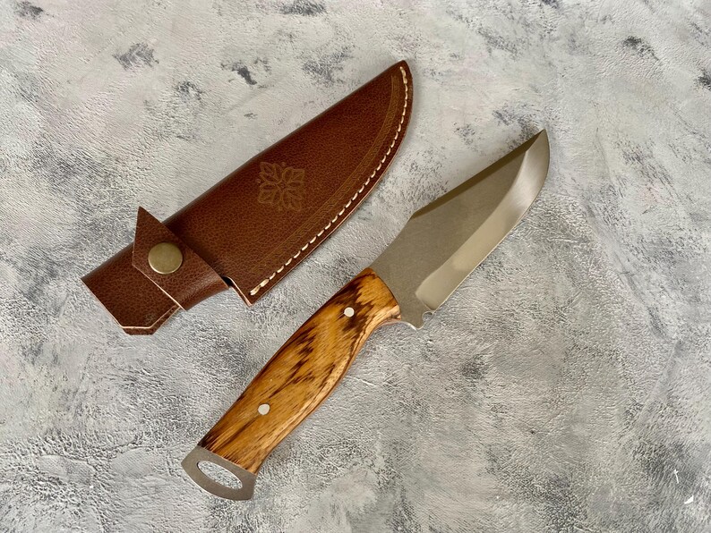 K&ouml;nnte beinhalten: Eine braune Lederscheide mit einem dekorativen Design und ein Messer mit Holzgriff und silberner Klinge.