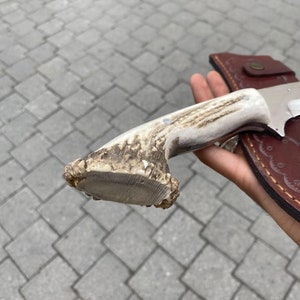 May include: A hunting knife with a bone handle and a leather sheath. The handle is made from a light-colored antler, and the blade is silver. The sheath is brown with a button closure.