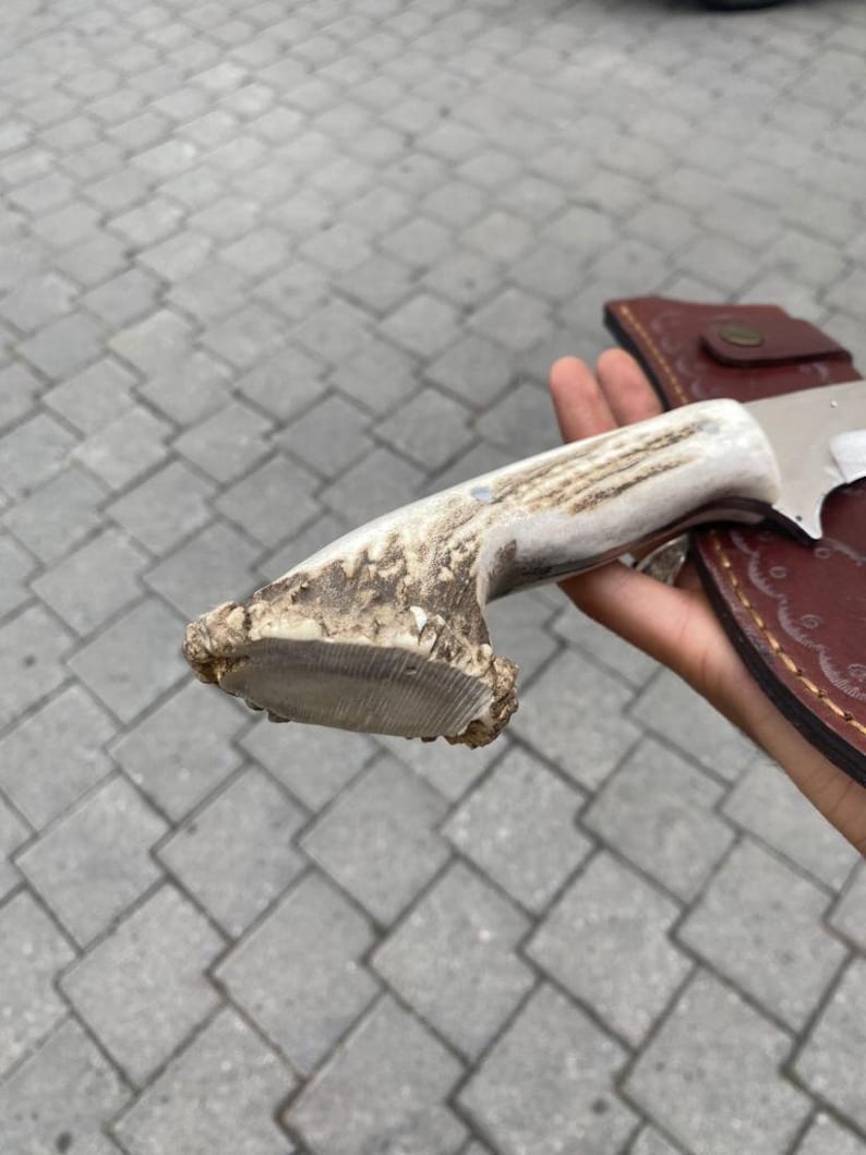 May include: A hunting knife with a bone handle and a leather sheath. The handle is made from a light-colored antler, and the blade is silver. The sheath is brown with a button closure.