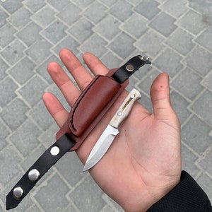May include: A small, handcrafted knife with a light-colored handle and a silver blade, accompanied by a brown leather sheath and a black leather strap with silver snap closures. The knife is designed for outdoor use.