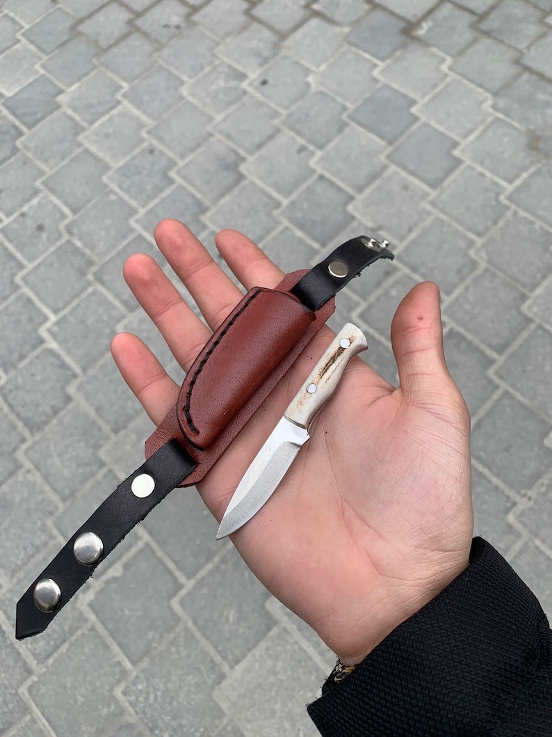 May include: A small, handcrafted knife with a light-colored handle and a silver blade, accompanied by a brown leather sheath and a black leather strap with silver snap closures. The knife is designed for outdoor use.