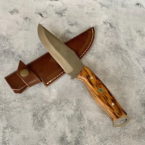 K&ouml;nnte beinhalten: Ein braunes Lederscheide mit einem silbernen Klingenmesser mit einem Holzgriff. Der Griff hat ein gr&uuml;nes und wei&szlig;es Design.