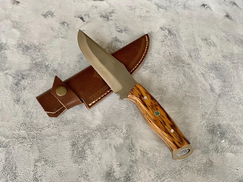 K&ouml;nnte beinhalten: Ein braunes Lederscheide mit einem silbernen Klingenmesser mit einem Holzgriff. Der Griff hat ein gr&uuml;nes und wei&szlig;es Design.