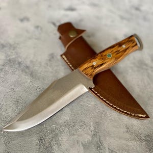K&ouml;nnte beinhalten: Ein Jagdmesser mit silberner Klinge und einem Holzgriff in einer braunen Lederscheide mit Naht.