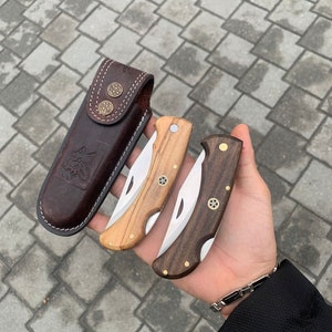 May include: A collection of three folding knives with wooden handles and stainless steel blades, alongside a brown leather sheath. The knives feature brass accents and are in varying shades of wood, from light to dark.