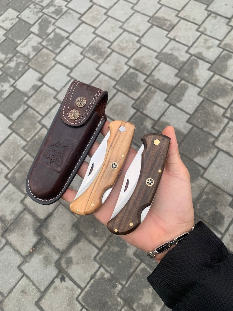 May include: A collection of three folding knives with wooden handles and stainless steel blades, alongside a brown leather sheath. The knives feature brass accents and are in varying shades of wood, from light to dark.