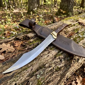 May include: A fixed-blade knife with a curved steel blade, a dark wood handle with brass accents, and a matching brown leather sheath featuring a snap closure and decorative stitching along the edges, presented as a complete set.
