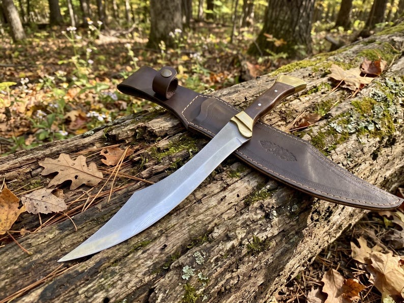 May include: A fixed-blade knife with a curved steel blade, a dark wood handle with brass accents, and a matching brown leather sheath featuring a snap closure and decorative stitching along the edges, presented as a complete set.