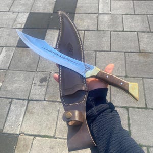 May include: A curved steel blade knife featuring an etched decorative pattern on the metal surface, a brown wooden handle with brass accents, and a matching brown leather sheath with visible stitching and a brass snap button closure.