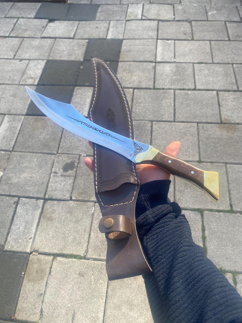 May include: A curved steel blade knife featuring an etched decorative pattern on the metal surface, a brown wooden handle with brass accents, and a matching brown leather sheath with visible stitching and a brass snap button closure.