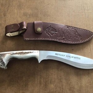 May include: A curved blade knife with a deer antler handle and a brown leather sheath. The blade has the text "MAJ ELOLF - KING OF BATTLE". The sheath has a decorative embossed design.