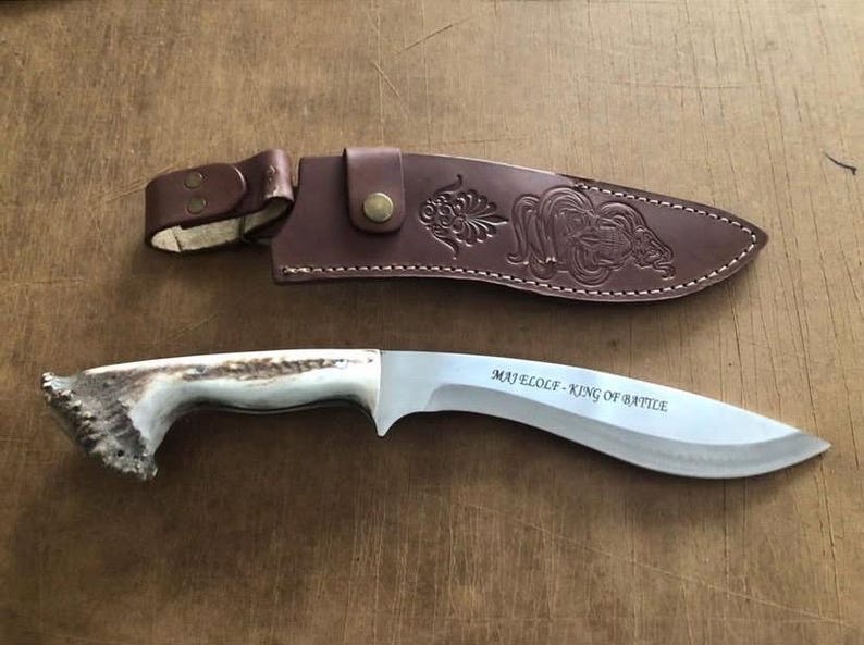 May include: A curved blade knife with a deer antler handle and a brown leather sheath. The blade has the text "MAJ ELOLF - KING OF BATTLE". The sheath has a decorative embossed design.