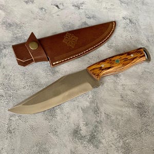 K&ouml;nnte beinhalten: Eine braune Lederscheide mit einem dekorativen Design und ein Messer mit Holzgriff und einer Klinge aus Edelstahl.