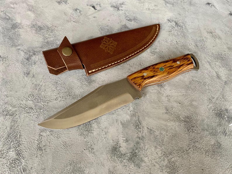 K&ouml;nnte beinhalten: Eine braune Lederscheide mit einem dekorativen Design und ein Messer mit Holzgriff und einer Klinge aus Edelstahl.