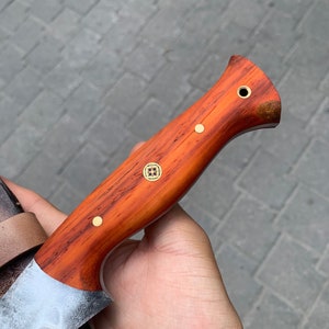 May include: A handcrafted knife with a wooden handle and a silver blade. The handle is a reddish-brown color with brass rivets and a decorative emblem. The blade has a slightly curved edge and a textured finish. The knife is partially visible.