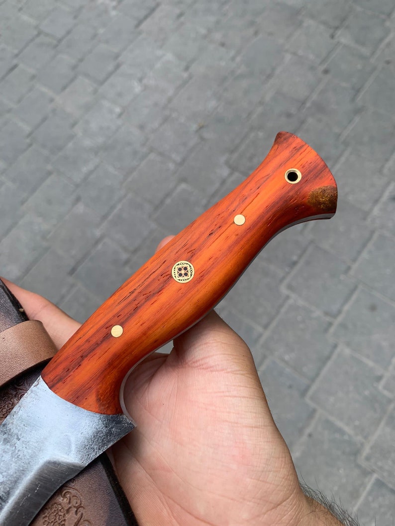 May include: A handcrafted knife with a wooden handle and a silver blade. The handle is a reddish-brown color with brass rivets and a decorative emblem. The blade has a slightly curved edge and a textured finish. The knife is partially visible.
