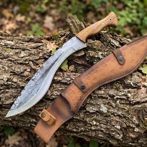 May include: A large, curved knife with a wooden handle and a leather sheath. The blade is silver and the handle is light brown. The sheath is brown and has a decorative pattern. The knife is resting on a log.