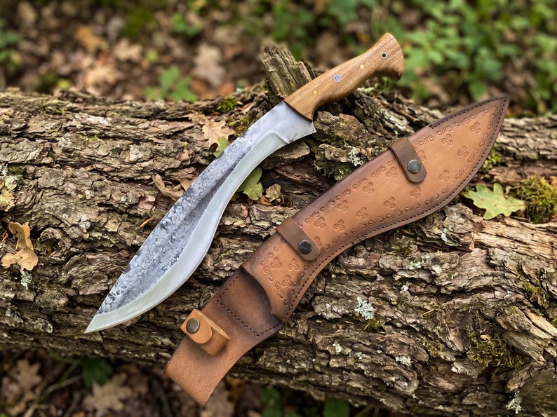 May include: A large, curved knife with a wooden handle and a leather sheath. The blade is silver and the handle is light brown. The sheath is brown and has a decorative pattern. The knife is resting on a log.