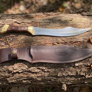 May include: A curved steel blade knife with a dark brown wooden handle, brass bolster, and pommel. Below the knife lies a matching dark brown leather sheath featuring decorative stitching, a snap button closure, and an embossed leaf pattern.