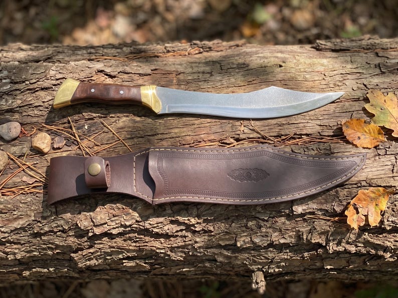 May include: A curved steel blade knife with a dark brown wooden handle, brass bolster, and pommel. Below the knife lies a matching dark brown leather sheath featuring decorative stitching, a snap button closure, and an embossed leaf pattern.