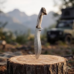 May include: A hunting knife with a curved blade and a light-colored antler handle is embedded in a tree stump. The knife is centered in the frame, with a blurred background of trees, mountains, and a vehicle.