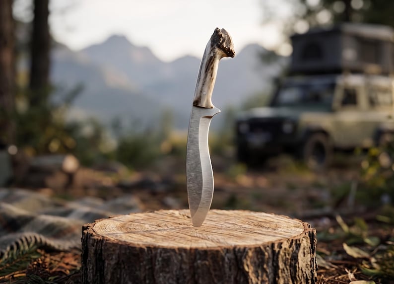 May include: A hunting knife with a curved blade and a light-colored antler handle is embedded in a tree stump. The knife is centered in the frame, with a blurred background of trees, mountains, and a vehicle.