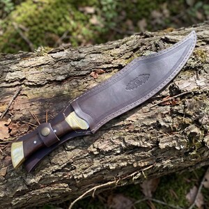 May include: A fixed-blade knife with a dark brown wooden handle and brass bolster, secured in a matching dark brown leather sheath. The sheath features decorative embossed border patterns and a central floral motif, with a snap button strap.