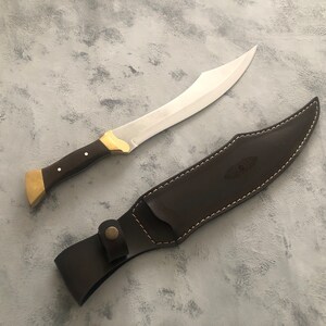May include: A curved steel blade knife with a dark brown wooden handle, brass bolster, and brass pommel. The knife is accompanied by a matching dark brown leather sheath featuring white contrast stitching and a snap button closure strap.