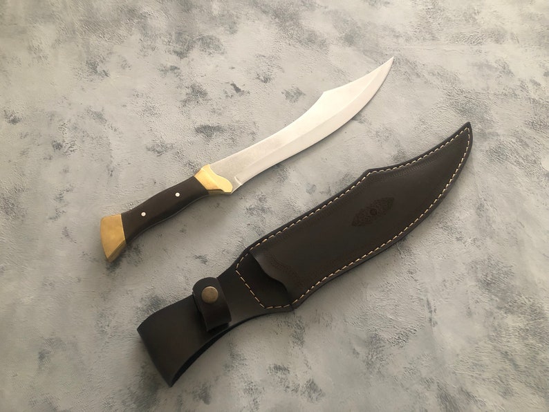 May include: A curved steel blade knife with a dark brown wooden handle, brass bolster, and brass pommel. The knife is accompanied by a matching dark brown leather sheath featuring white contrast stitching and a snap button closure strap.