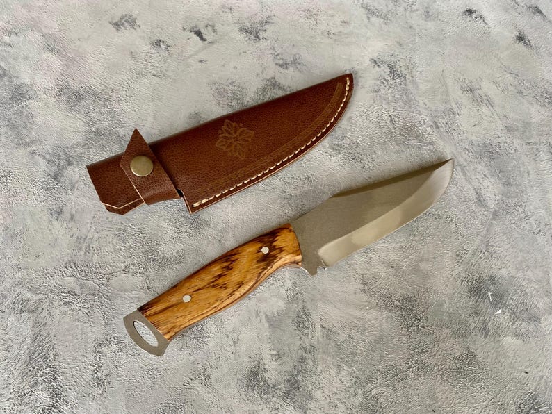 K&ouml;nnte beinhalten: Eine braune Ledertasche f&uuml;r ein Messer mit einem dekorativen Design und ein Messer mit Holzgriff und silberner Klinge.