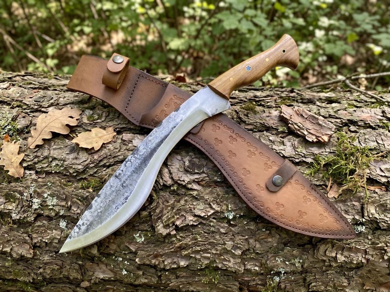 May include: A large, curved knife with a wooden handle and a leather sheath. The blade has a textured finish. The sheath is brown leather with decorative embossed patterns. The knife and sheath are resting on a tree trunk.