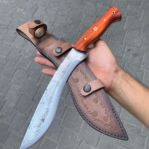 May include: A large, curved blade knife with a wooden handle and a leather sheath. The knife has a silver blade and a brown handle with brass rivets. The sheath is brown leather with decorative floral embossing and a belt loop.