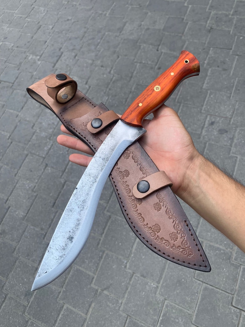May include: A large, curved blade knife with a wooden handle and a leather sheath. The knife has a silver blade and a brown handle with brass rivets. The sheath is brown leather with decorative floral embossing and a belt loop.