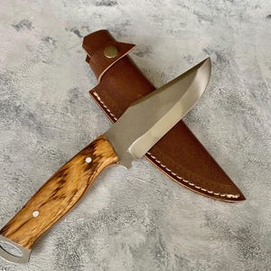 K&ouml;nnte beinhalten: Ein Jagdmesser mit silberner Klinge und einem Holzgriff in einer braunen Lederscheide mit Naht.