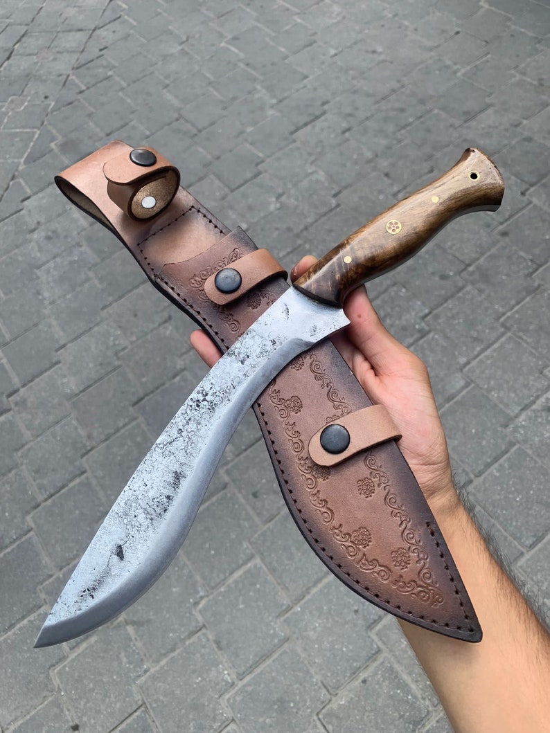 May include: A large, curved knife with a wooden handle and a leather sheath. The blade has a textured, aged appearance. The sheath is brown with decorative embossed patterns and a leather strap with a button closure. The handle has brass accents.